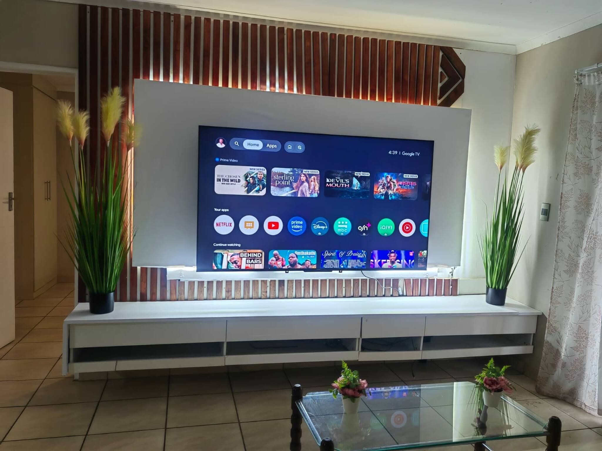 A modern living room with a large flat-screen TV mounted on a wooden wall, a glass coffee table, and potted plants.