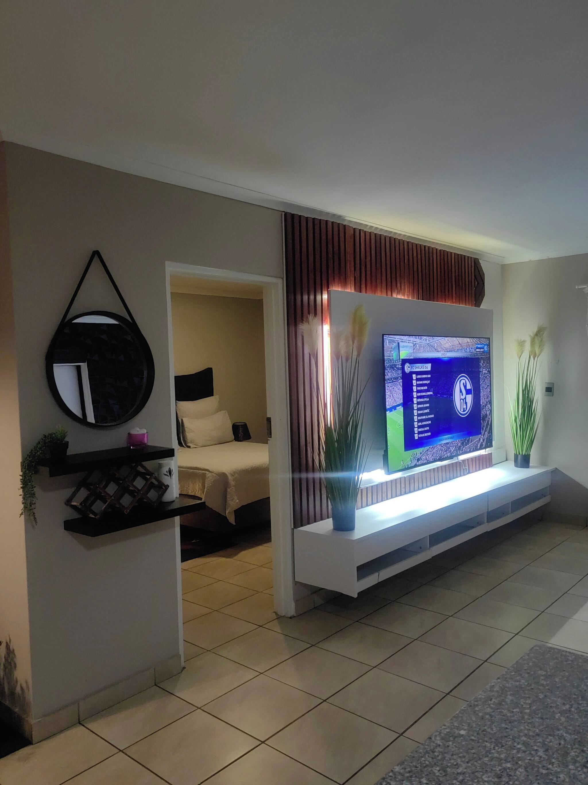 A modern living room with a flat-screen TV mounted on a white console, a bed with a white headboard, and a potted plant on the console.