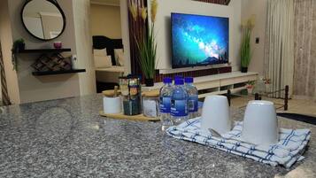 A modern living room with a flat-screen TV, a kitchen counter with water bottles, and a dining area with a placemat.