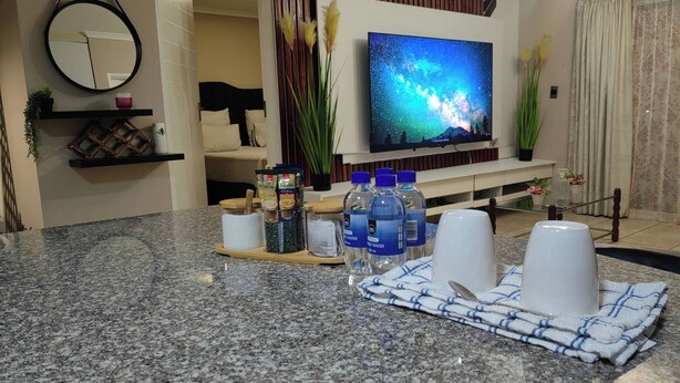 Uma sala de estar moderna com televisão de tela plana, bancada de cozinha com garrafas de água e área de jantar com jogo americano.