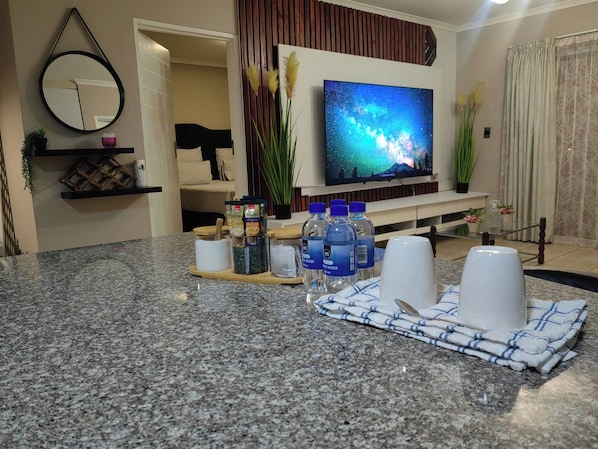 A modern living room with a flat-screen TV, a kitchen counter with water bottles, and a dining area with a placemat. - Luxury Boutique Apartment in Secure Pretoria Estate (Pretoria)