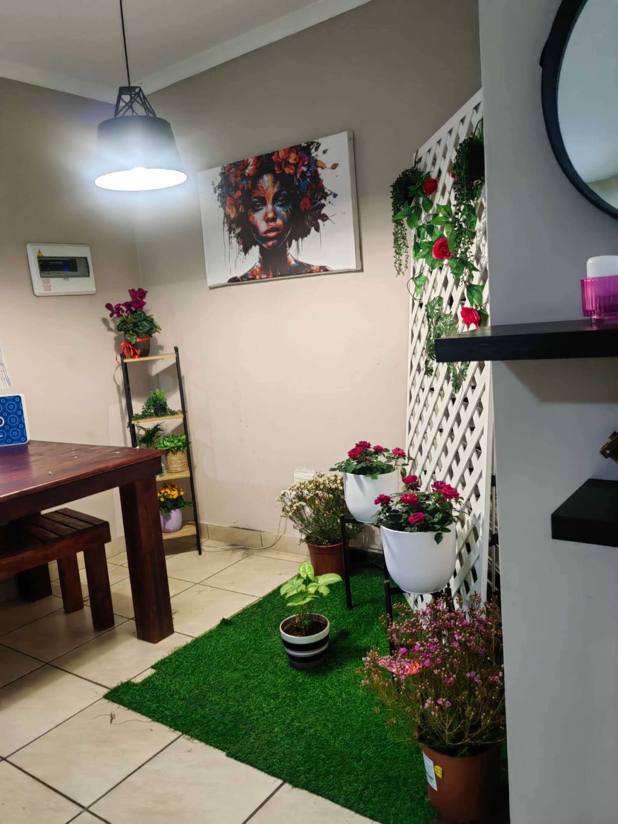Un comedor con una mesa de madera, sillas, una lámpara colgante, un cuadro en la pared, plantas en macetas y un felpudo de césped artificial verde.
