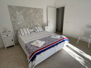 Un dormitorio con cama, mesitas de noche, una silla y una puerta que da a otra habitación.