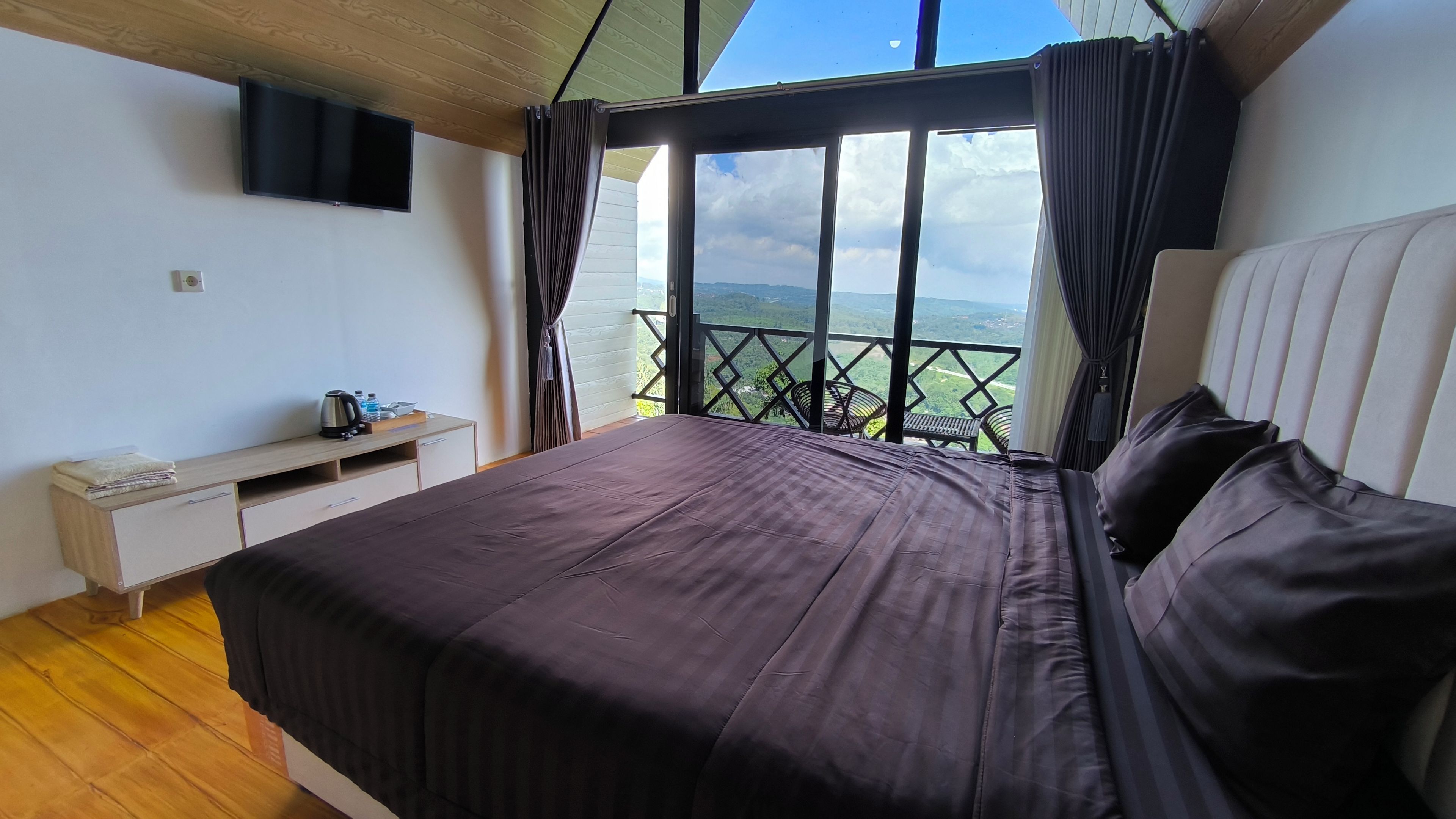 A bedroom with a bed, a TV mounted on the wall, a desk with a kettle, and a view of the outdoors.