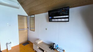 A room with a wooden ceiling, a mounted TV displaying various apps, a small desk with a kettle and water bottles, and a wooden door. - Senja Kita Glamping (Salatiga)