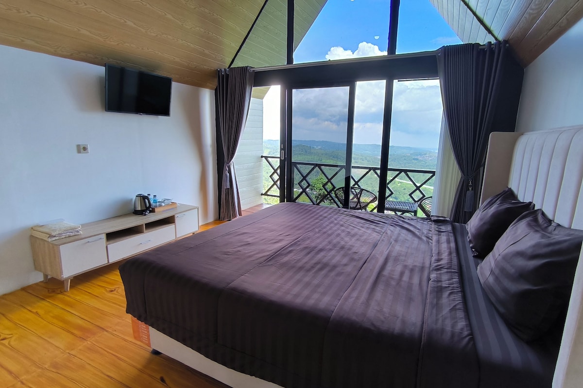 Un dormitorio con cama, televisor, suelo de madera, ventana con cortinas y vistas al paisaje.