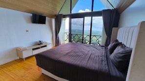 Un dormitorio con cama, televisor, suelo de madera, ventana con cortinas y vistas al paisaje.