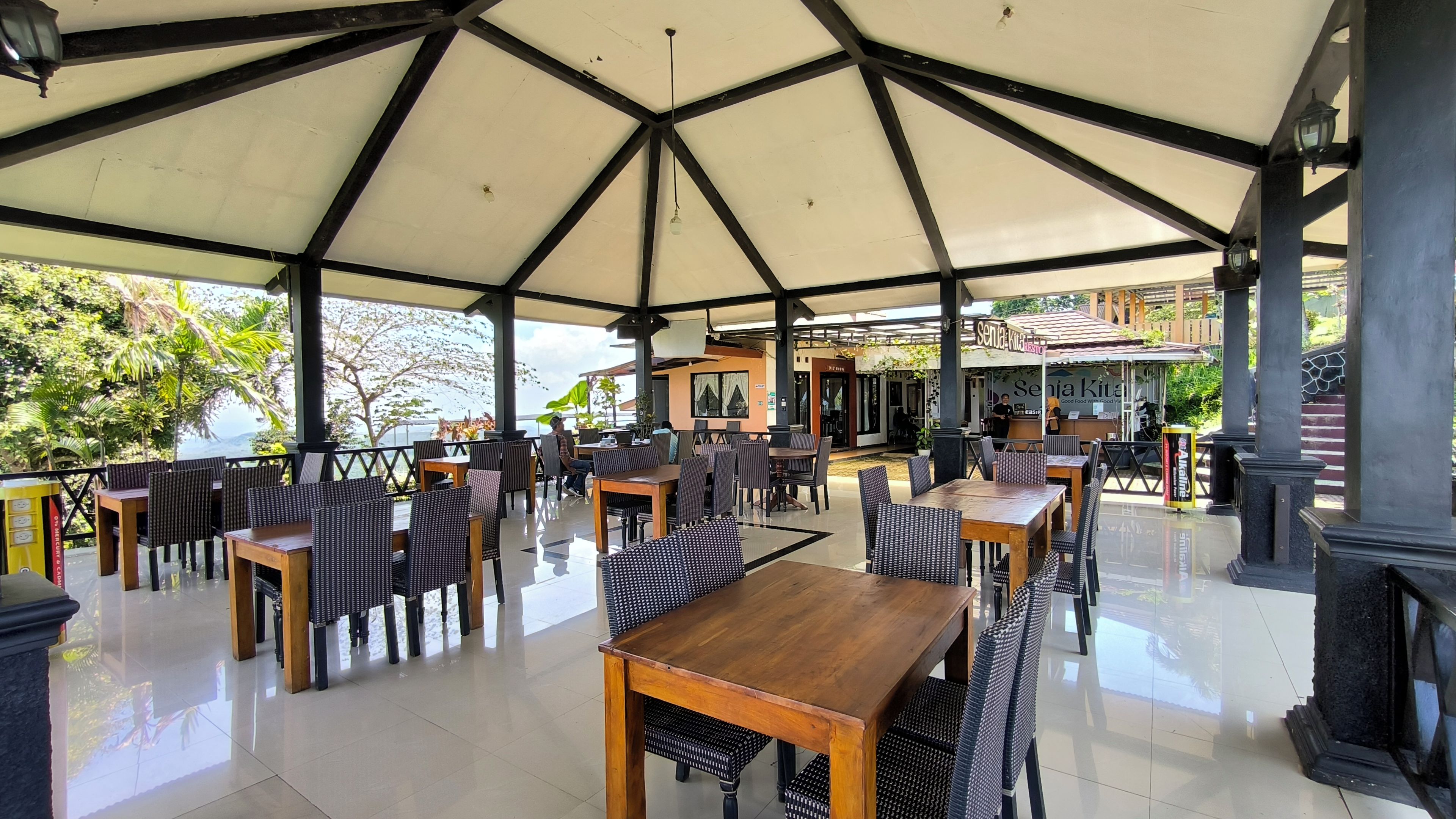 An outdoor cafe with tables and chairs, a covered area, and a view of the sea.