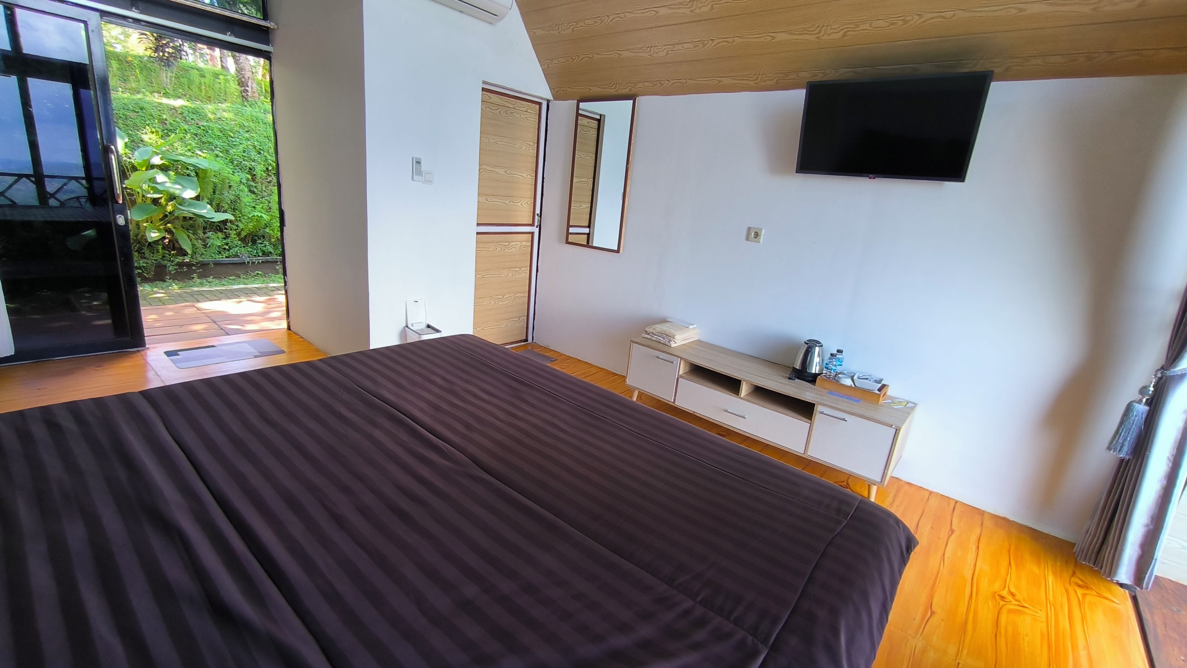 A bedroom with a large bed, a TV mounted on the wall, and a small desk with a kettle and a bottle.