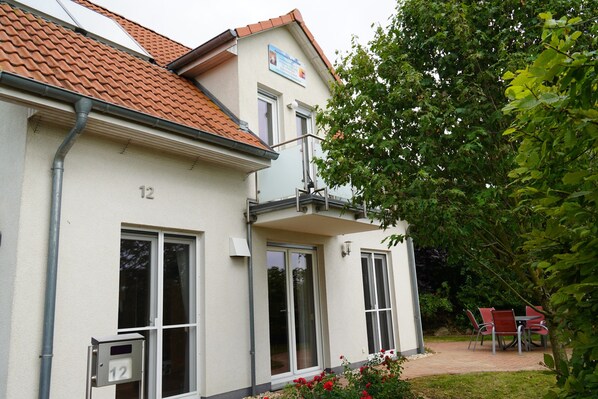 A two-story building with a tiled roof, numbered 12, featuring a balcony and a small garden area with red flowers.
