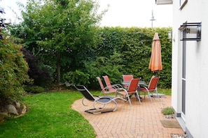 A patio with a table and chairs, surrounded by a hedge and a lawn.