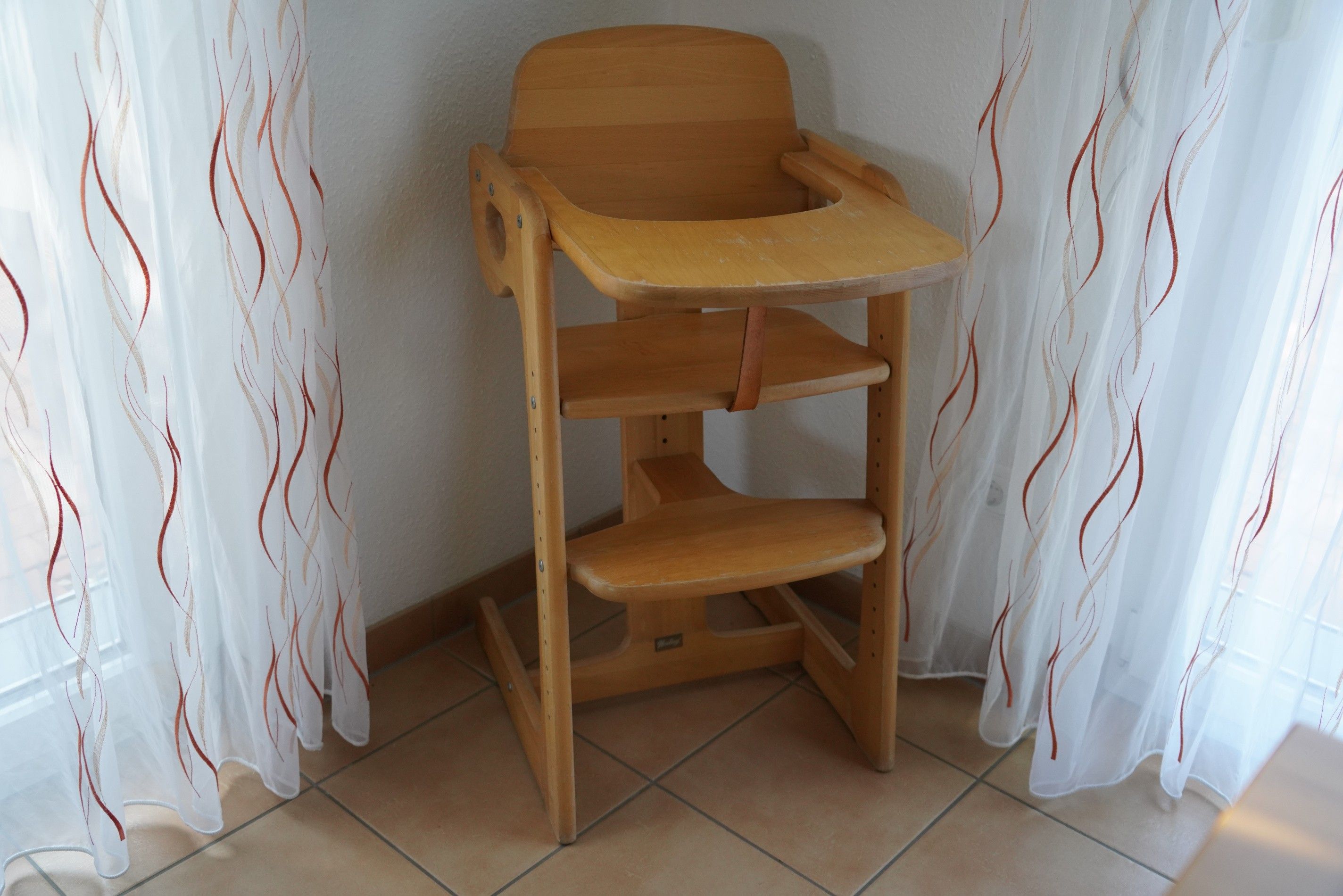 Ein hölzerner Hochstuhl mit Tablett vor einer Wand mit durchsichtigen Vorhängen.