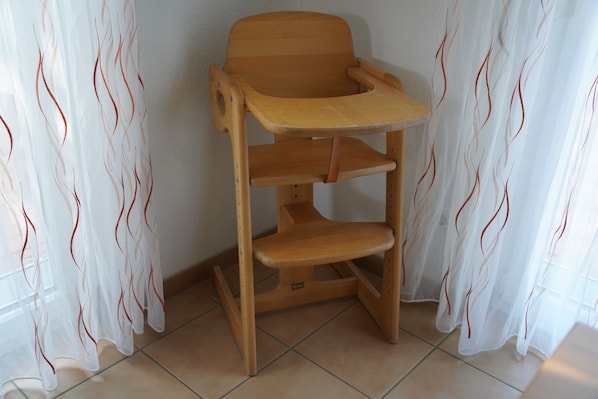 A wooden high chair with a tray table against a wall with sheer curtains.