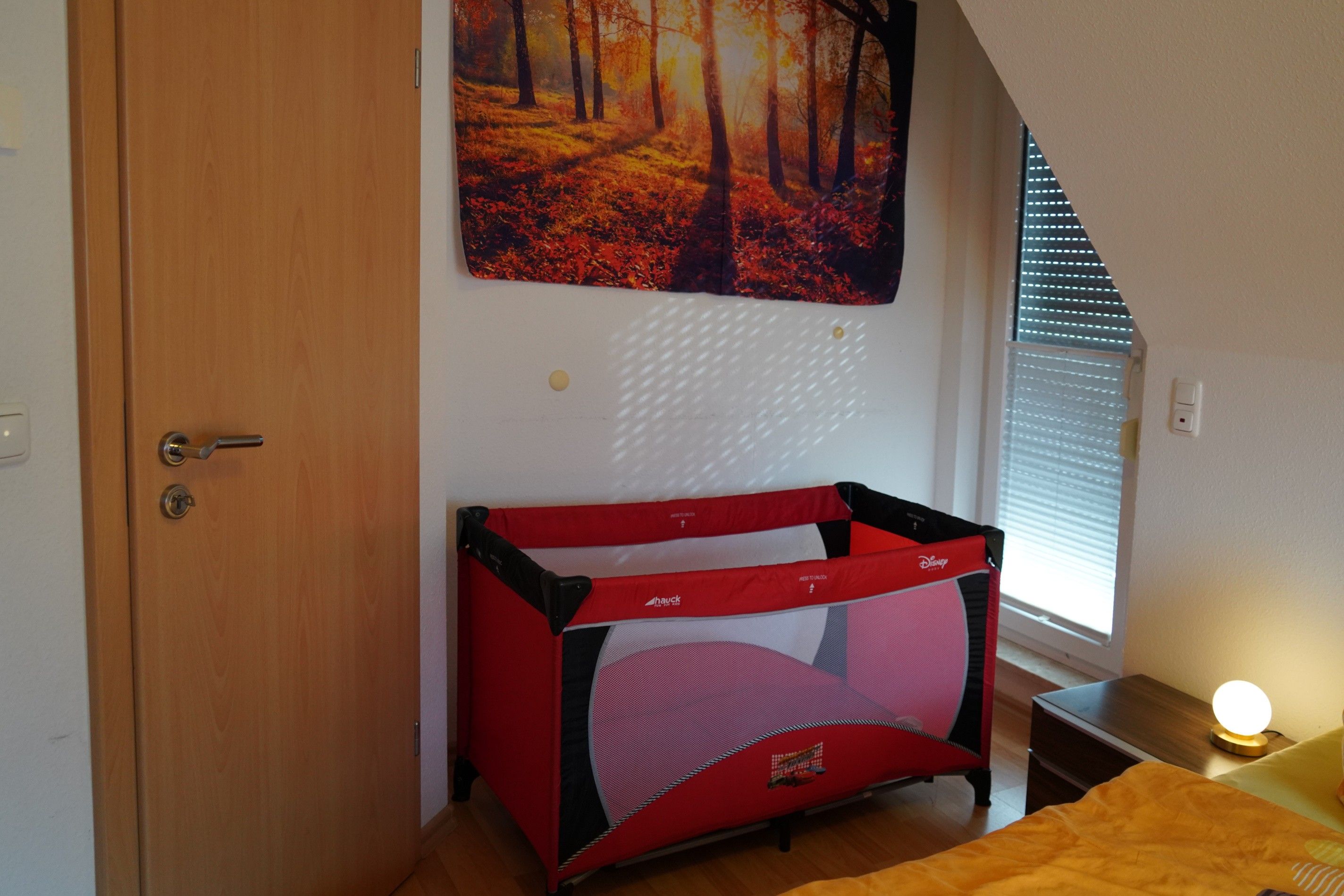Ein Zimmer mit Holztür, einem roten und schwarzen Babybett, einem Waldgemälde und einem Fenster mit Jalousien.