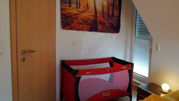 Ein Zimmer mit Holztür, einem roten und schwarzen Babybett, einem Waldgemälde und einem Fenster mit Jalousien.