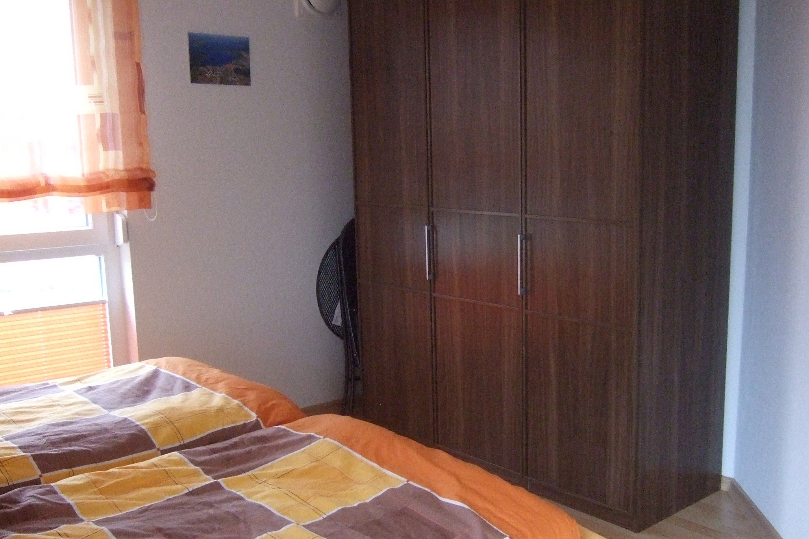 Ein Schlafzimmer mit einem Bett, einem Kleiderschrank, einem Wandventilator und einem Fenster mit Vorhängen.