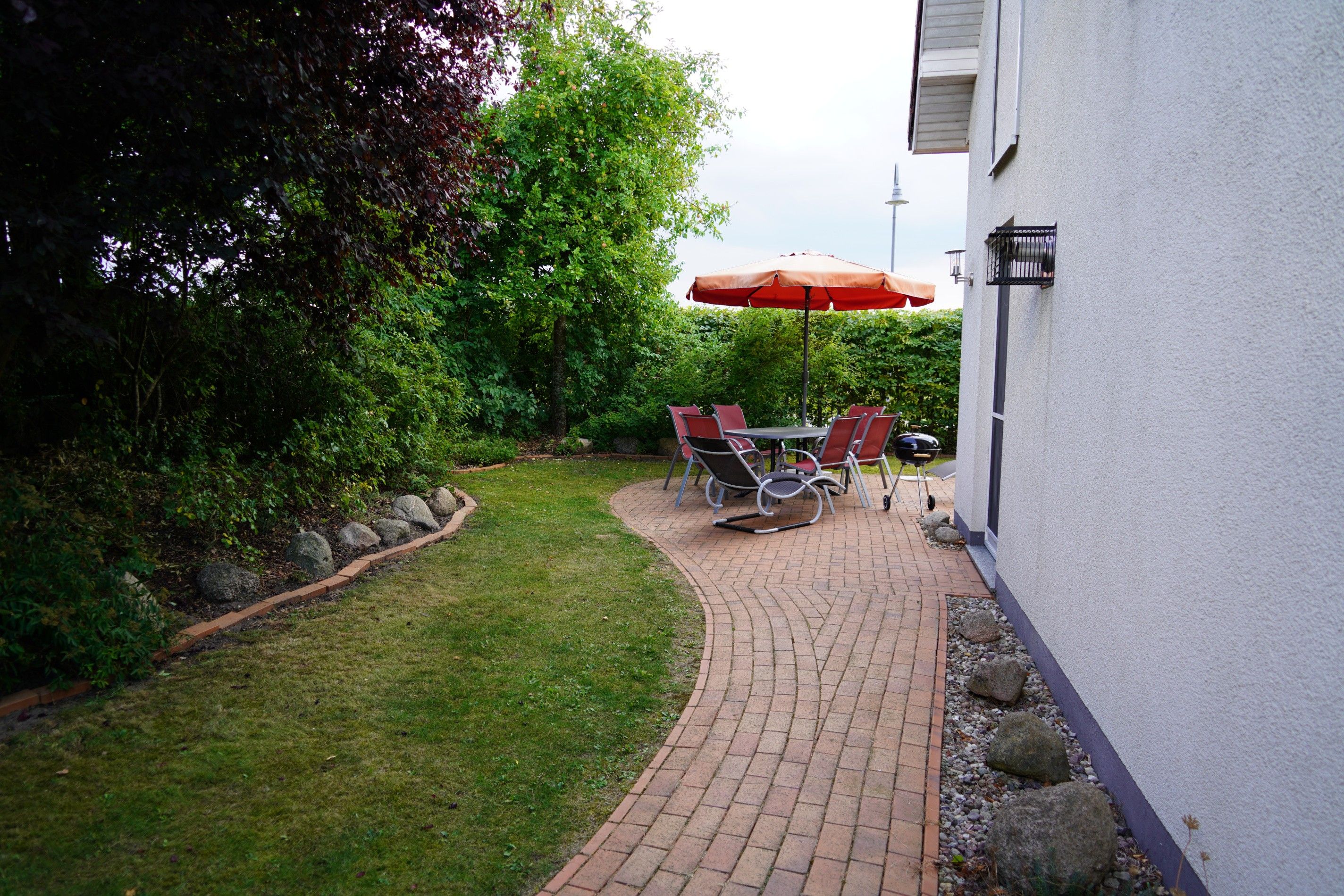 Eine gepflasterte Terrasse mit Tisch, Stühlen, einem roten Sonnenschirm und einem Grill.