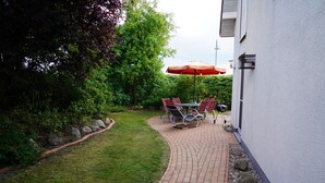 Um pátio pavimentado com mesa, cadeiras, um guarda-sol vermelho e uma churrasqueira.