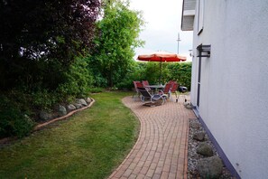 A paved patio with a table and chairs, a red umbrella, and a grill.