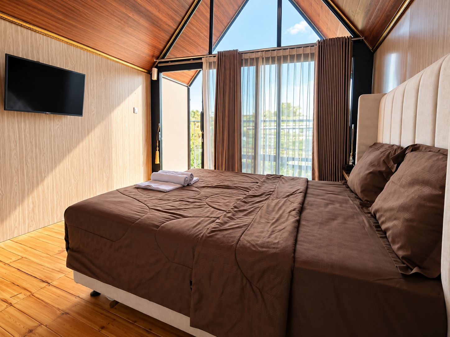 A bedroom with a large bed, a flat-screen TV mounted on the wall, and a window with curtains.