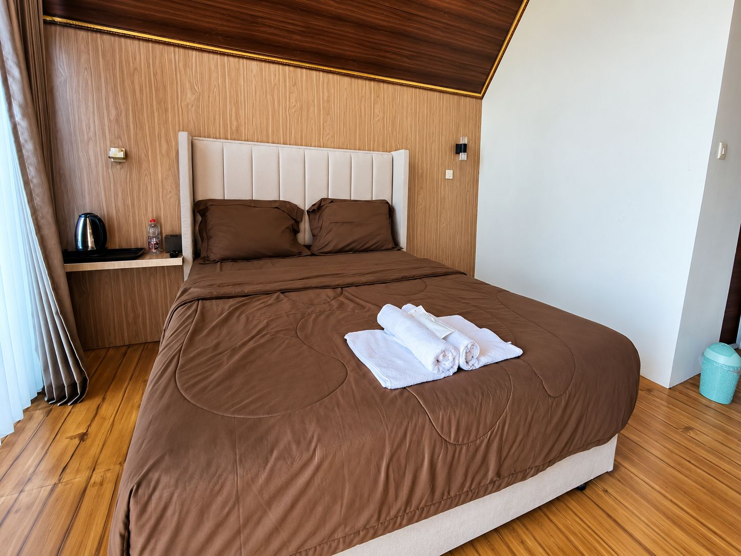 A hotel room with a wooden headboard, a bed with brown bedding, a small bedside table with a kettle and a bottle, and a rolled towel on the bed.