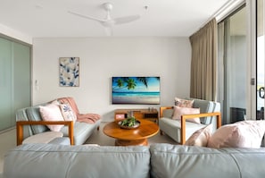 A modern living room with a round coffee table, two sofas, a flat-screen TV, and a ceiling fan. - Belle Escapes 1104 Harbour Lights Top Floor Ocean & City Views (Cairns City)