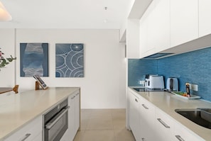 A modern kitchen with white cabinetry, a blue backsplash, and two abstract wall art pieces. - Belle Escapes 1104 Harbour Lights Top Floor Ocean & City Views (Cairns City)