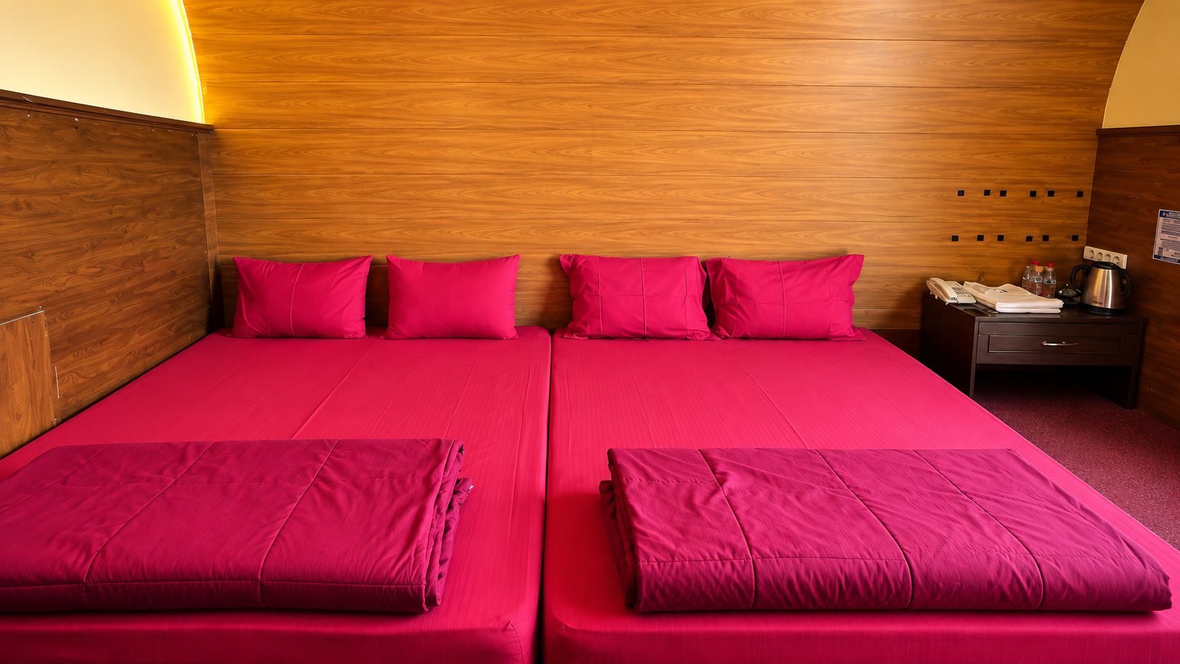 Una cama doble con ropa de cama roja, un cabecero de madera y una mesita de noche con un teléfono y una tetera.