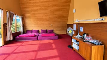 Una habitación con techo inclinado de madera, alfombra roja, dos camas moradas con almohadas a juego, un escritorio de madera con un ventilador y una ventana con cortinas.