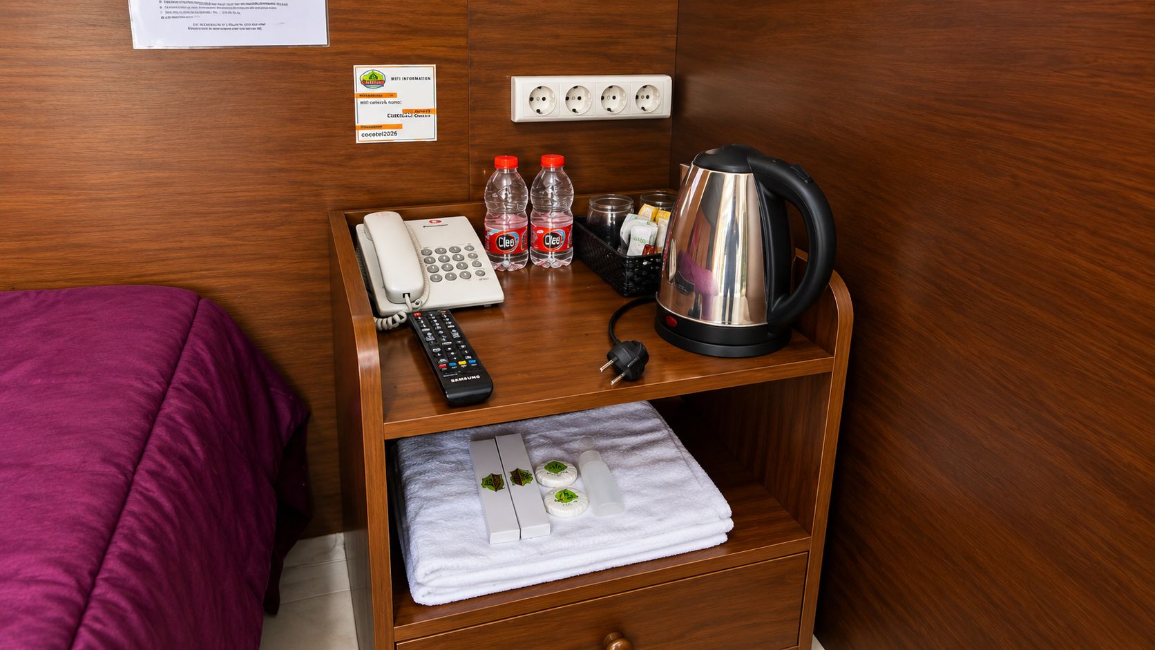 A wooden nightstand with a phone, remote control, and a kettle.