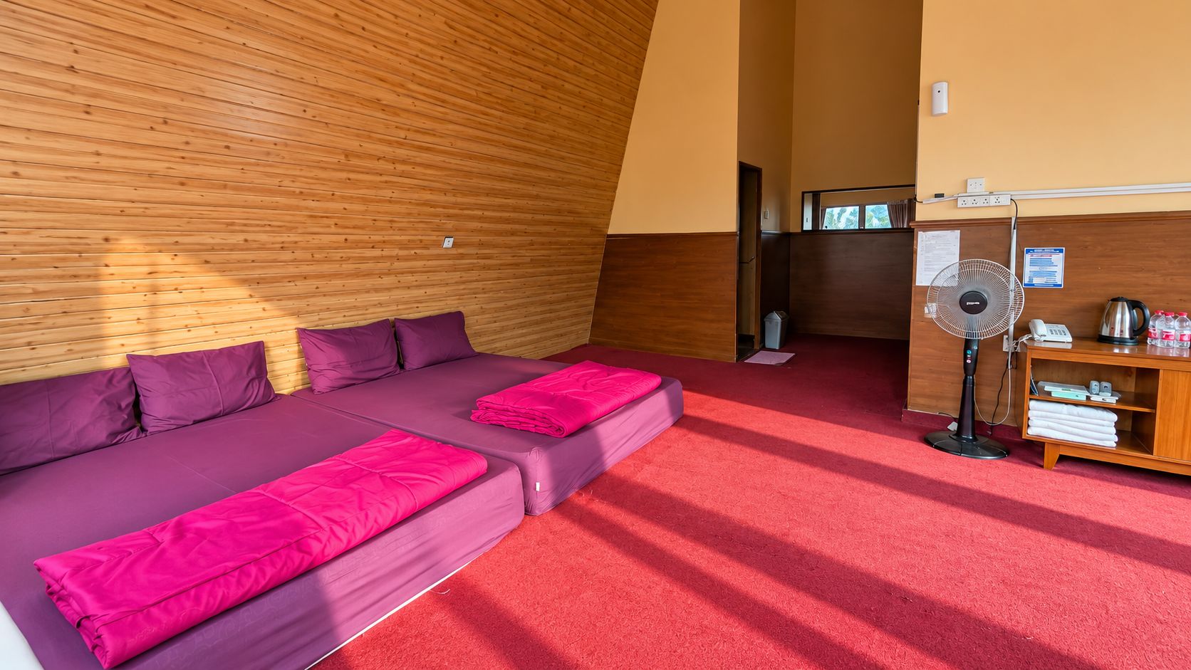 Una habitación con techo de madera inclinado, alfombra roja, sofá con ropa de cama morada, un ventilador de pie y una estantería con objetos.