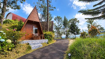 A small, triangular wooden cabin with a stone staircase leading to the entrance, surrounded by lush greenery and trees.