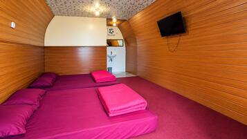 Una habitación con paredes de madera, una cama con ropa de cama rosa, un lavamanos pequeño blanco y un televisor montado en la pared.
