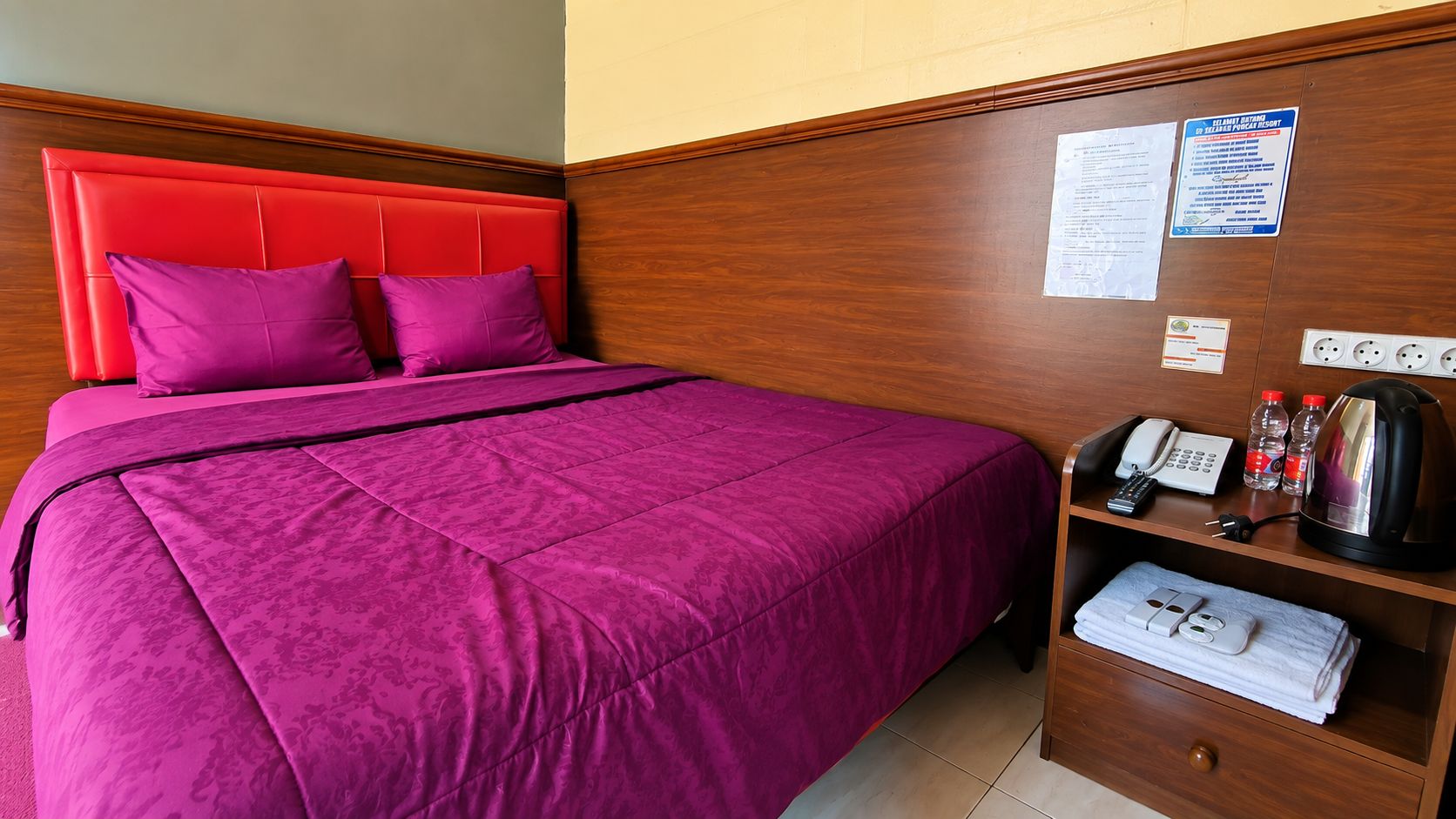 A hotel room with a bed, a wooden headboard, a bedside table with a phone, a kettle, and a folded towel.