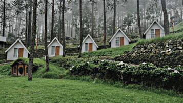 A row of small, white cabins with wooden shutters, surrounded by a forested area with tall trees and a stone wall.