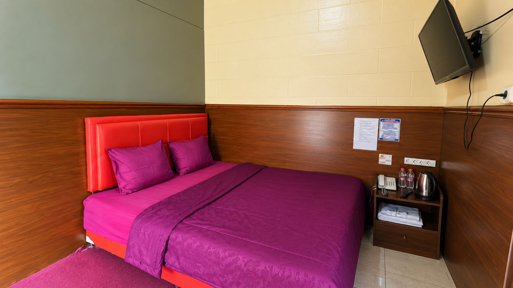 Habitación de hotel con cama, cabecero de madera, estante pequeño con artículos de aseo y televisor montado en la pared.