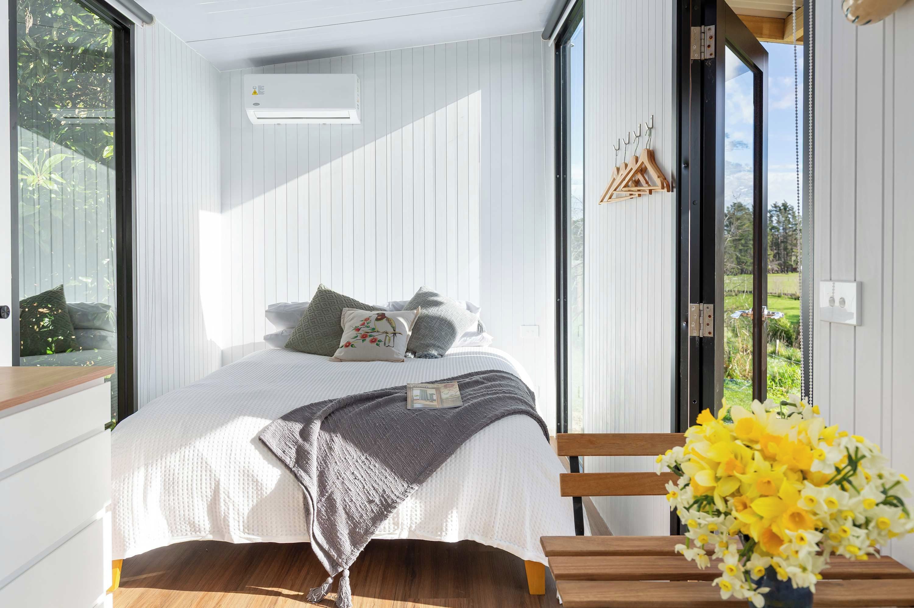 A bedroom with a bed, pillows, a bedside table, a chair, and a window with a view of greenery.