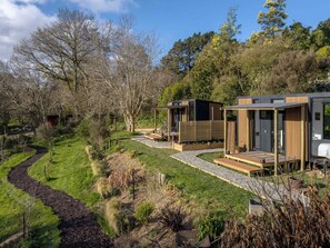 A modern outdoor living area with a pathway leading to a wooden deck, surrounded by lush greenery and trees. - Papa Ora Healthy Earth Stays X by Tiny Away (Rangitūmau)