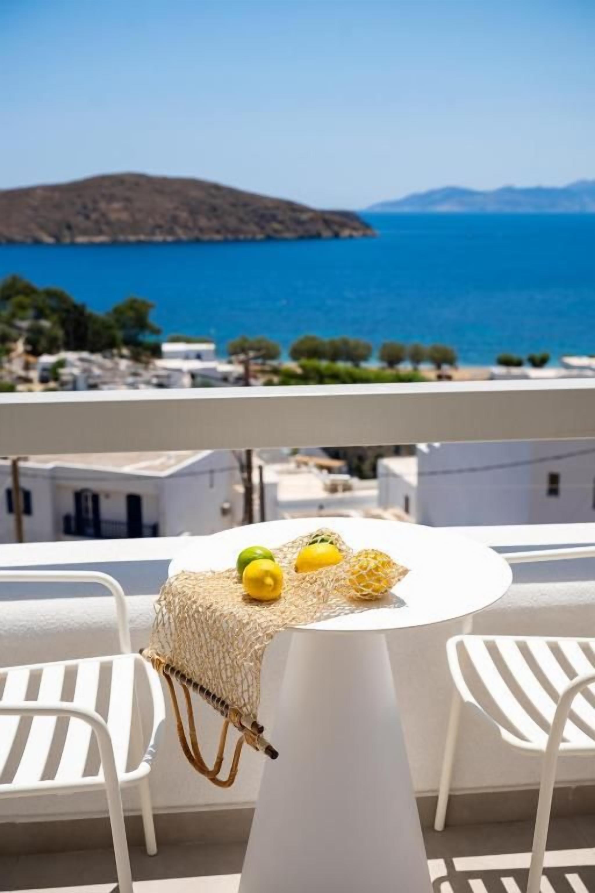 A balcony with a white table and chairs, overlooking a coastal view with buildings and a clear sky.