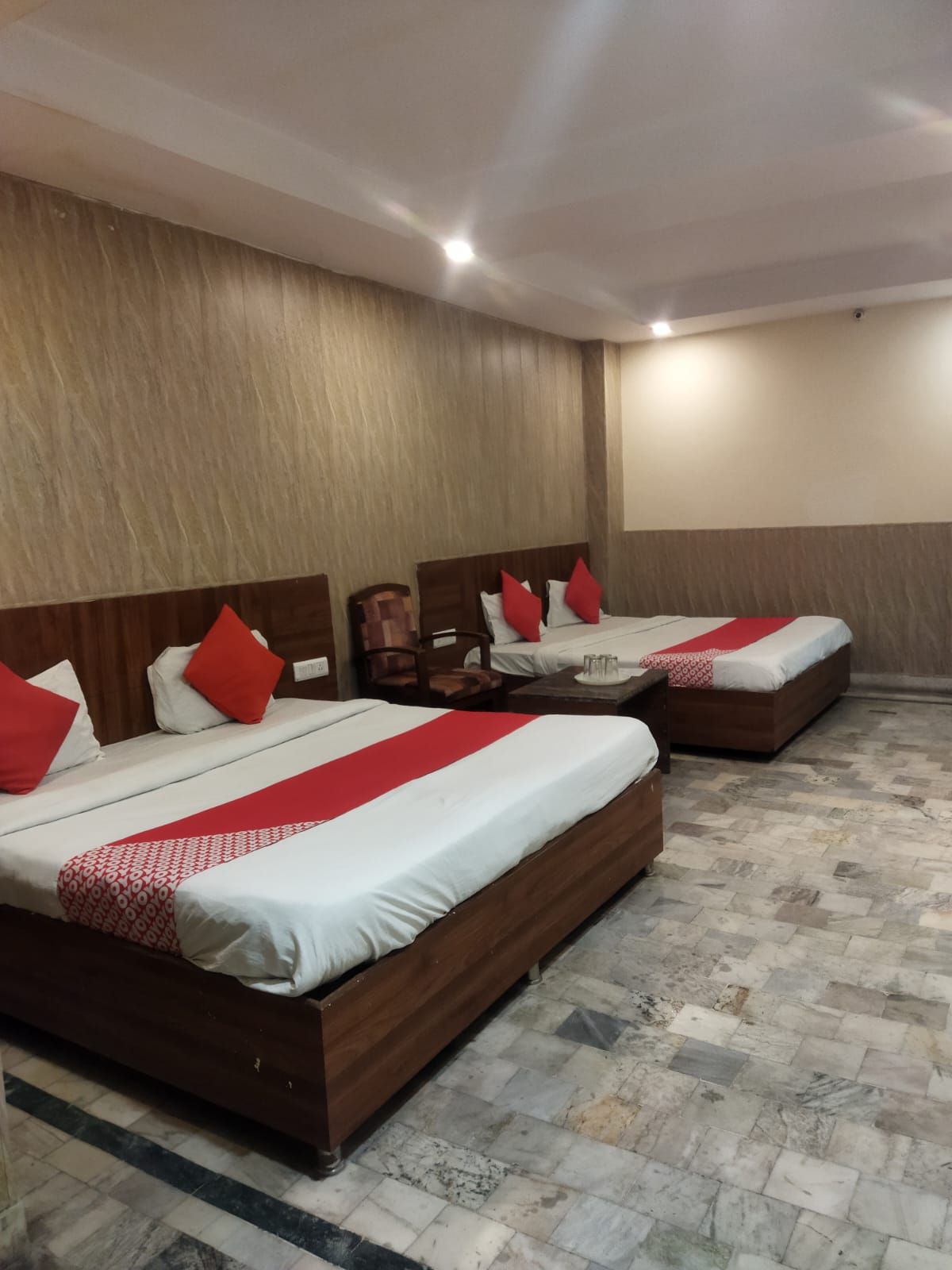 Habitación de hotel con dos camas, cada una con cabecero de madera, ropa de cama blanca y roja, y una mesita de noche con lámpara.