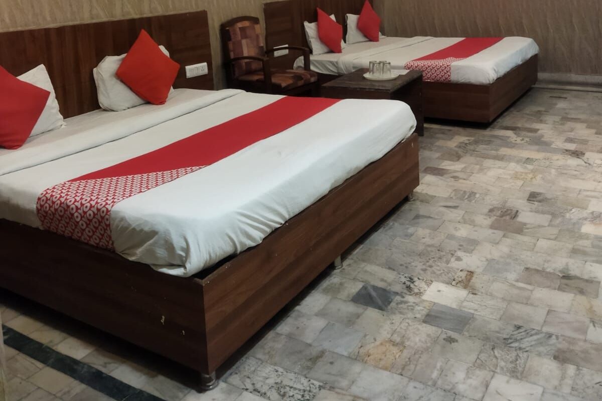 Quarto de hotel com duas camas, cada uma com cabeceira de madeira, roupa de cama vermelha e branca, e uma pequena mesa de cabeceira com abajur.