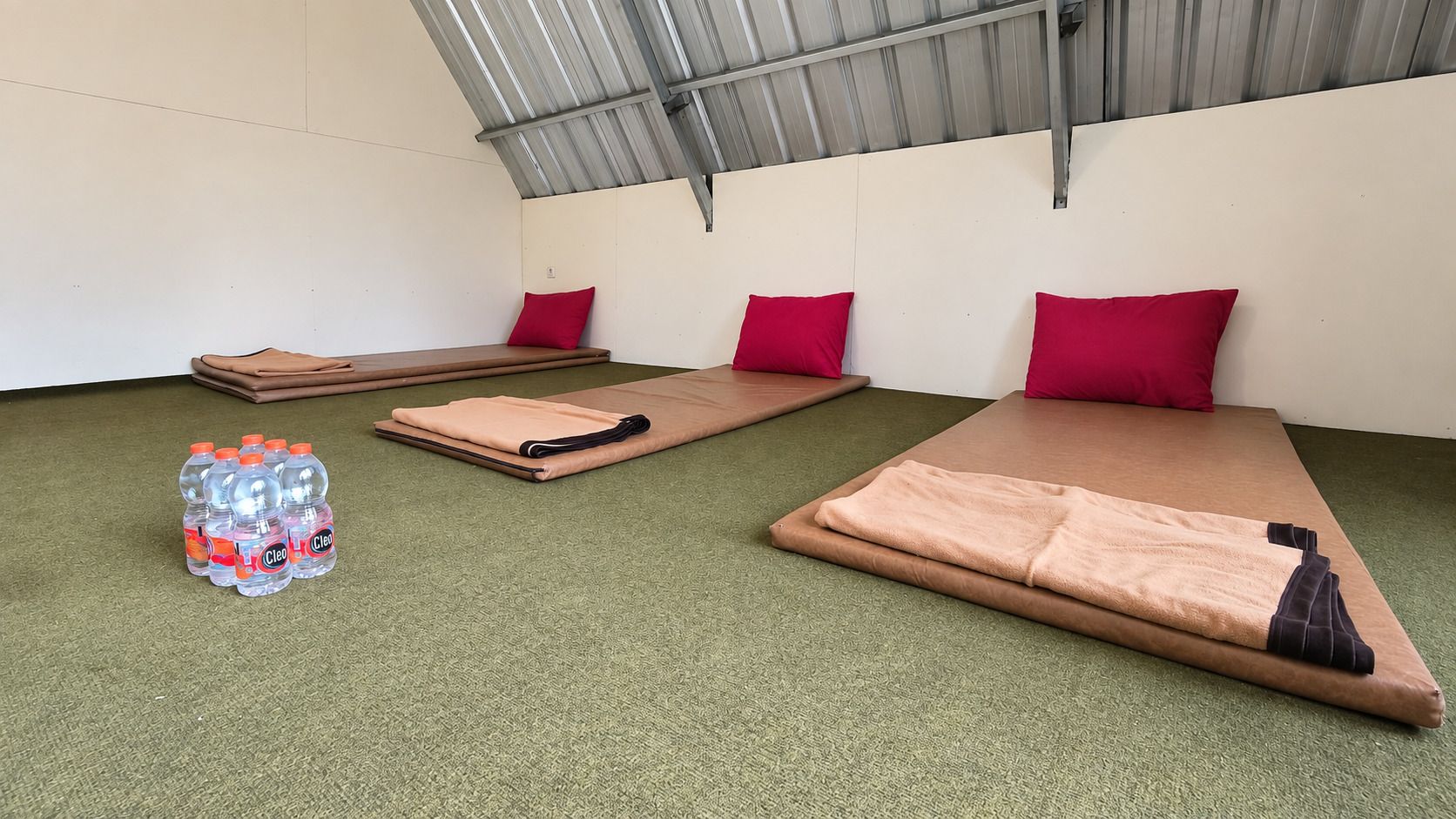 A room with three mats, each with a red pillow, and a stack of bottled water on the floor.