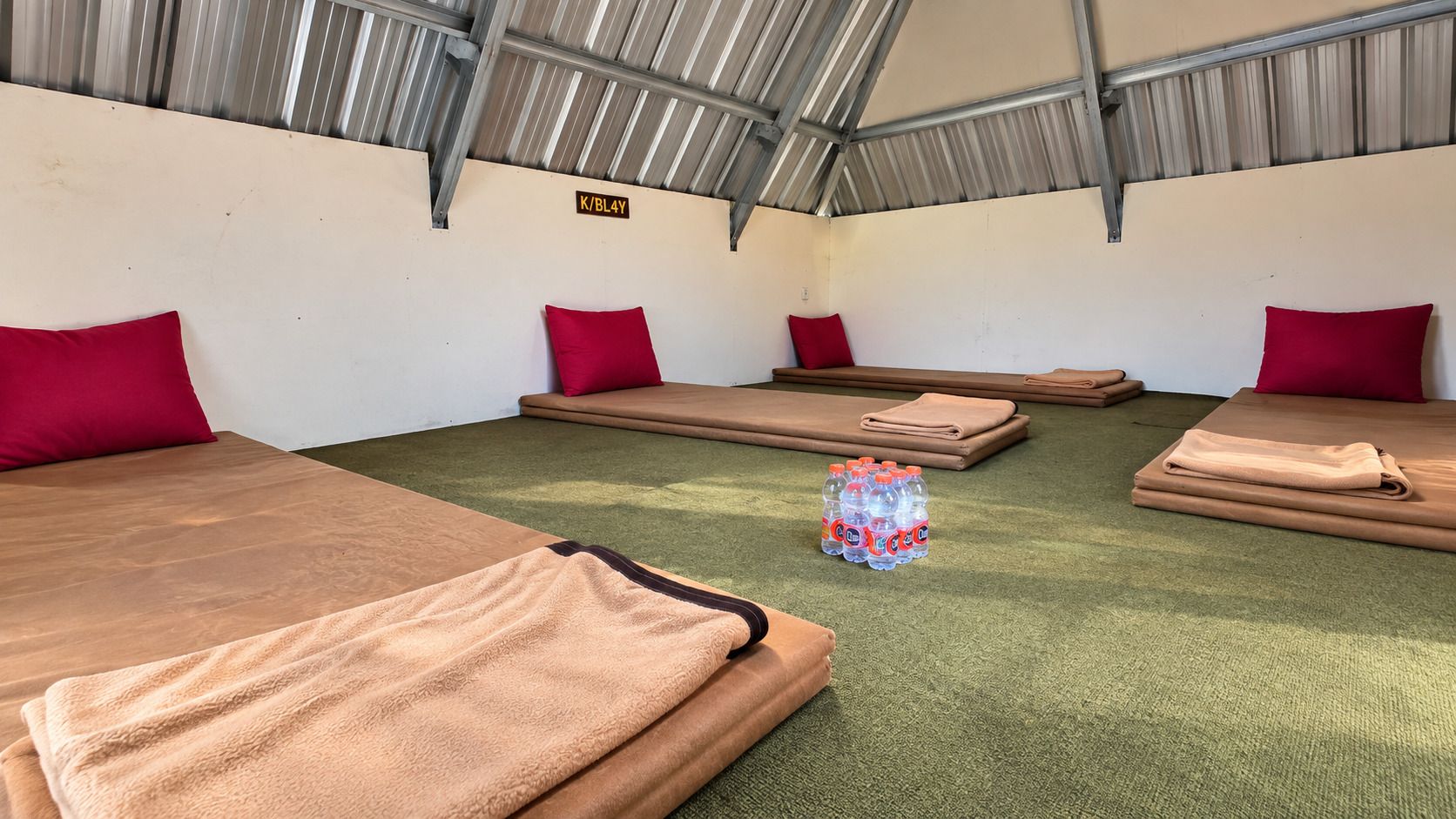 A room with four beds, each with a red pillow and a brown blanket. There is a stack of folded blankets and a pile of bottled water on the floor.