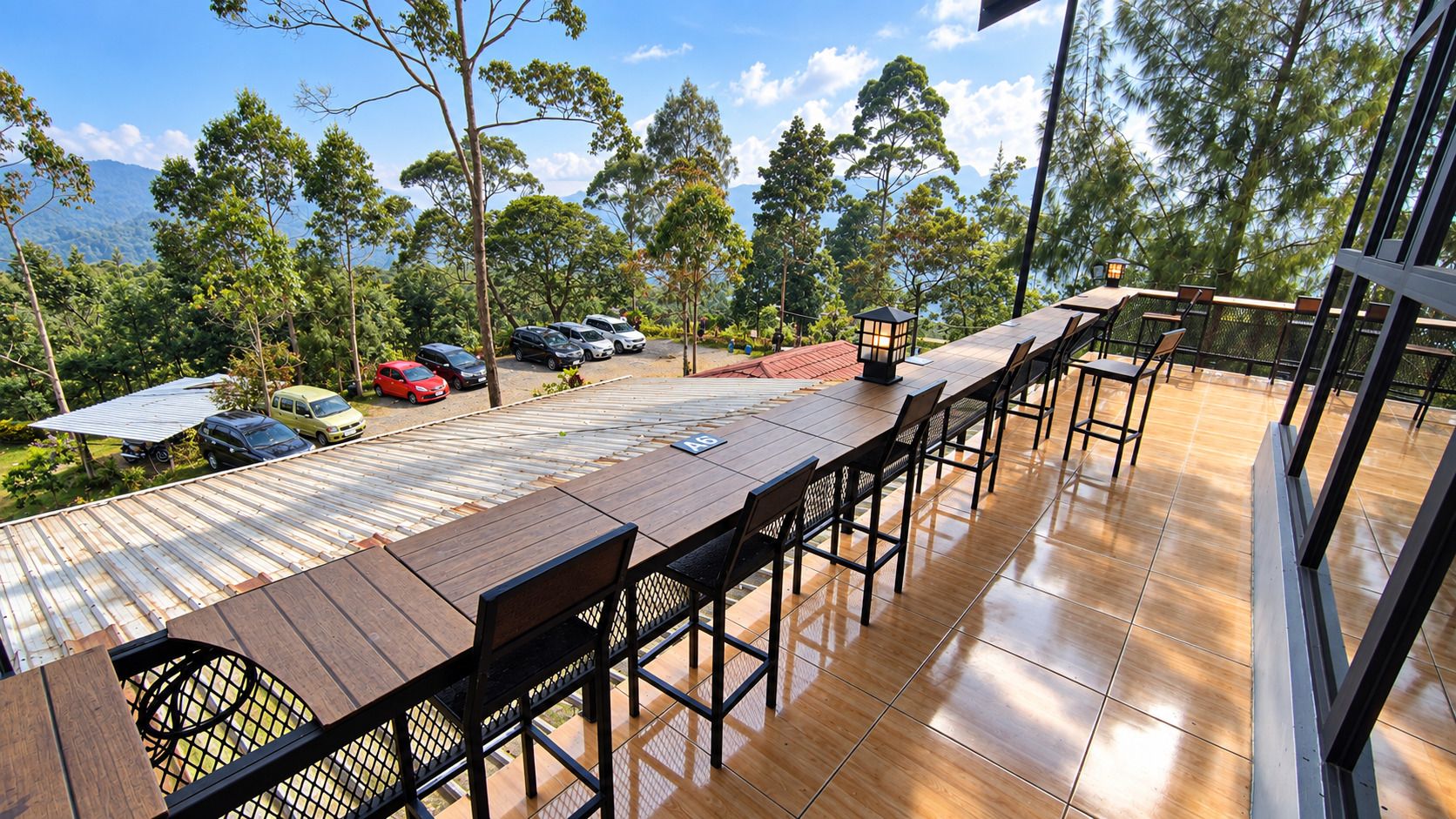 An outdoor dining area with a long wooden table and chairs, surrounded by a glass railing, overlooking a parking area with several cars and a mountainous backdrop.