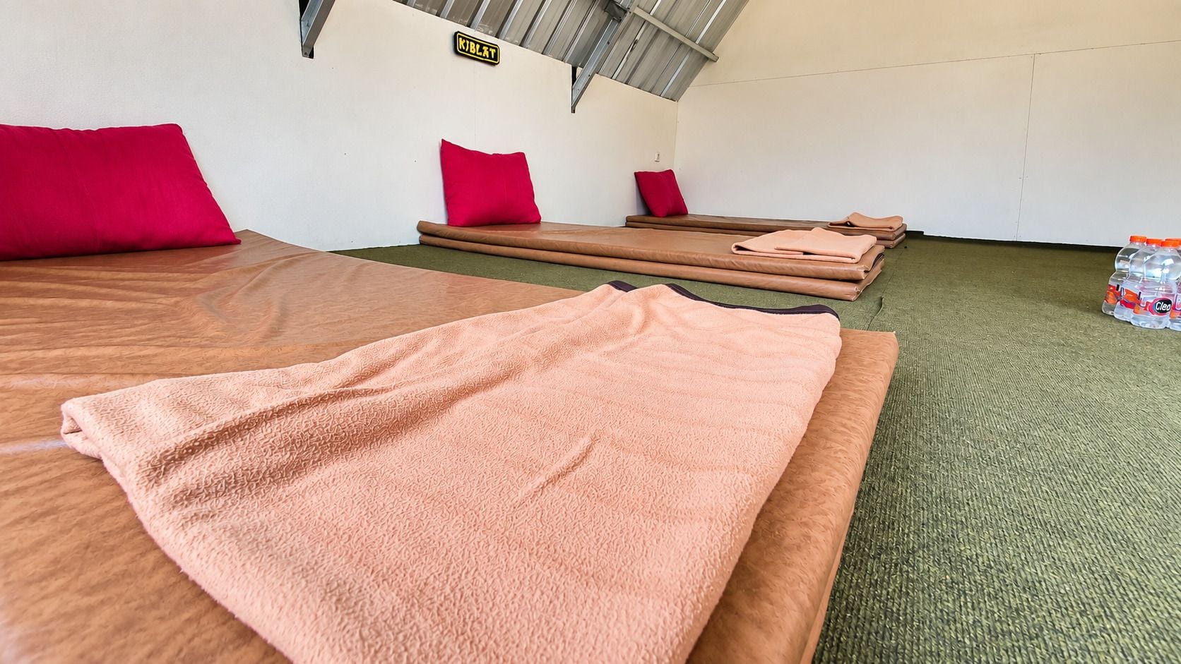 Quarto com duas camas, cada uma com uma toalha rosa e um travesseiro vermelho. Há duas pias de madeira com almofadas. Uma garrafa de água está visível no chão.
