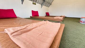 Quarto com duas camas, cada uma com uma toalha rosa e um travesseiro vermelho. Há duas pias de madeira com almofadas. Uma garrafa de água está visível no chão.