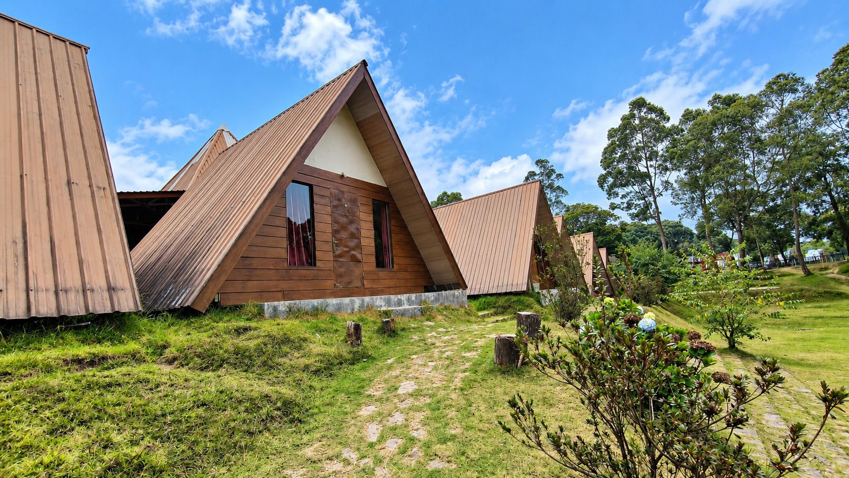 Una estructura de madera triangular con ventana y puerta visibles, rodeada de césped y árboles.