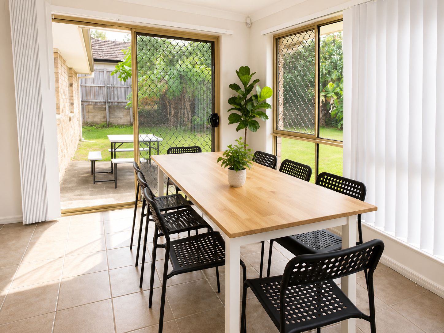 A dining area with a wooden table and black chairs, a potted plant, and a view of a backyard through a screened door.