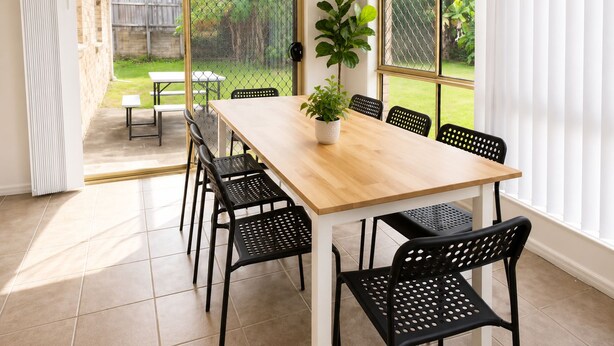 A dining area with a wooden table and black chairs, a potted plant, and a view of a backyard through a screened door. - 4 BR Family Retreat – 15 min to Ipswich (Redbank Plains)