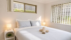 A neatly made bed with a rolled towel, two bedside lamps, a small plant on a nightstand, and a window with a metal screen. - 4 BR Family Retreat – 15 min to Ipswich (Redbank Plains)